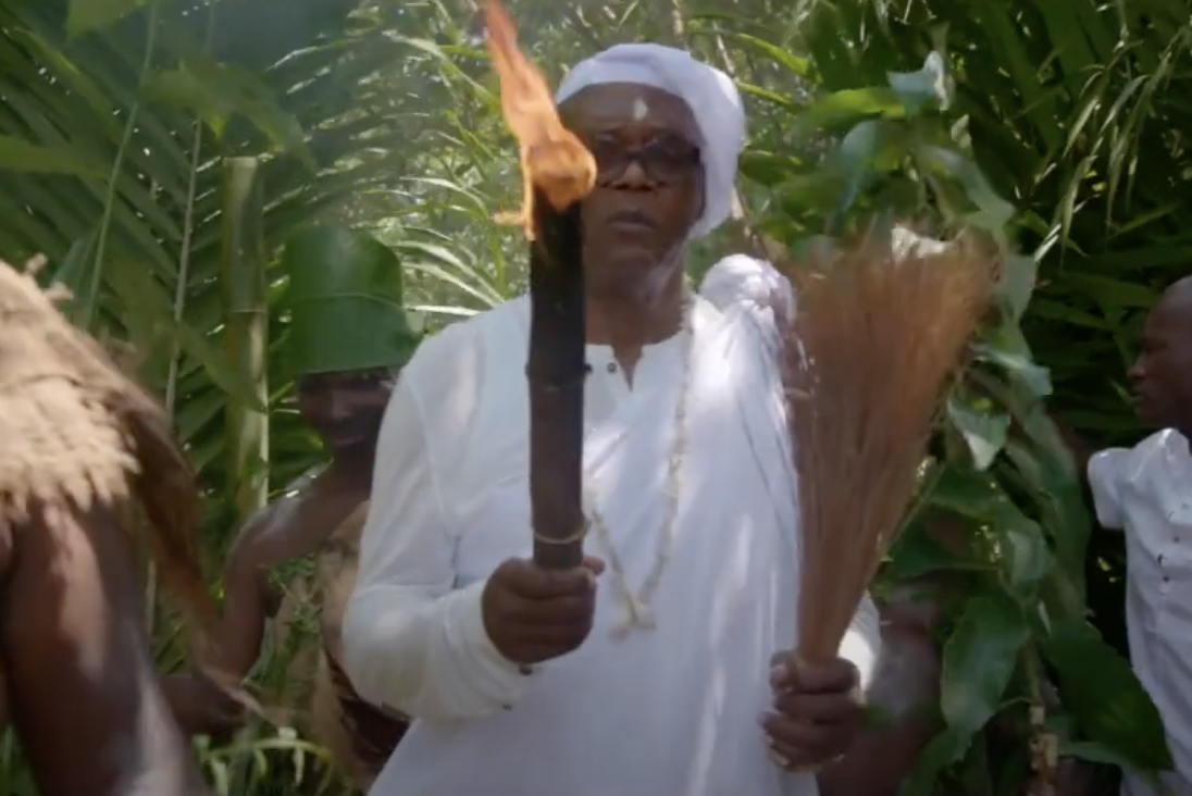 Samuel L. Jackson, dressed all in white, emerges from palm fronds holding a burning stick
