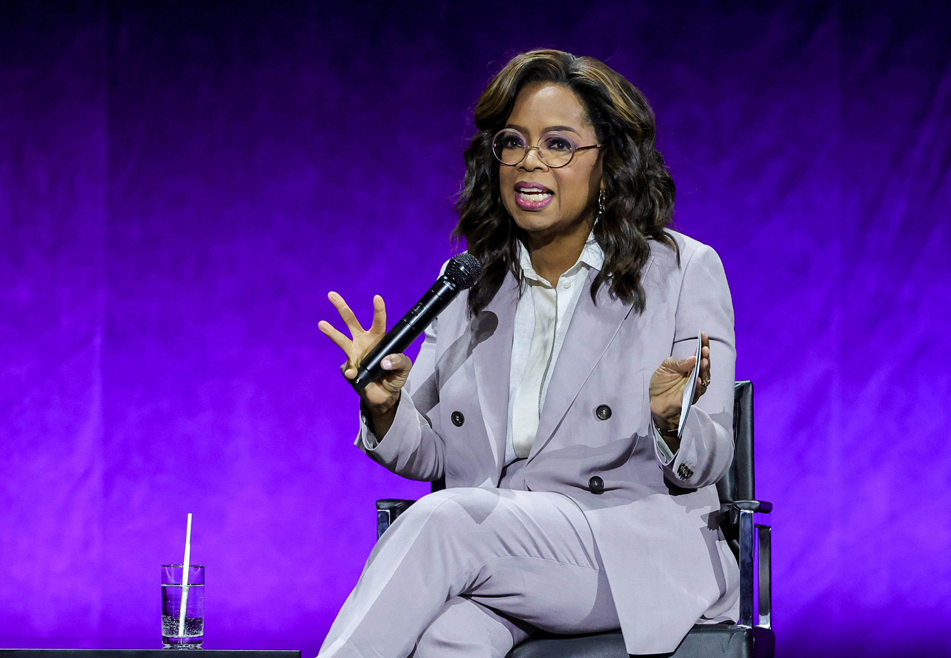 Oprah sitting on a stage in a chair, wearing a grey suit, speaking into a microphone