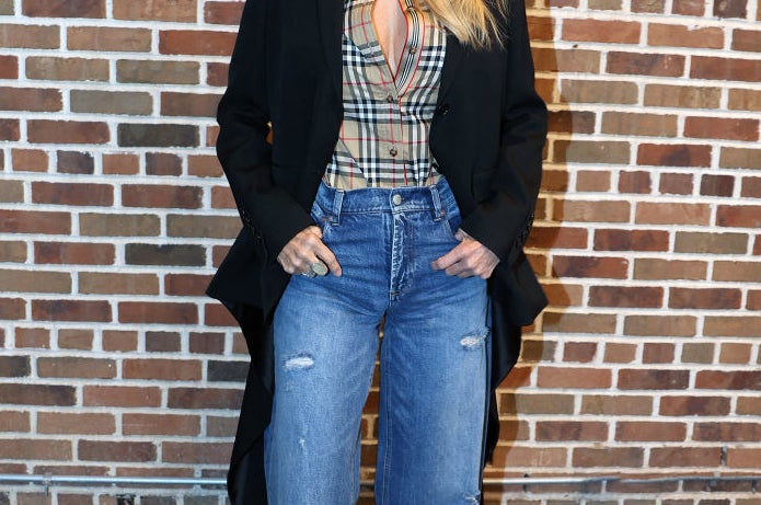 A closeup of Christie smiling as she poses for the camera against a brick wall. She&#x27;s wearing a plaid blouse and jeans