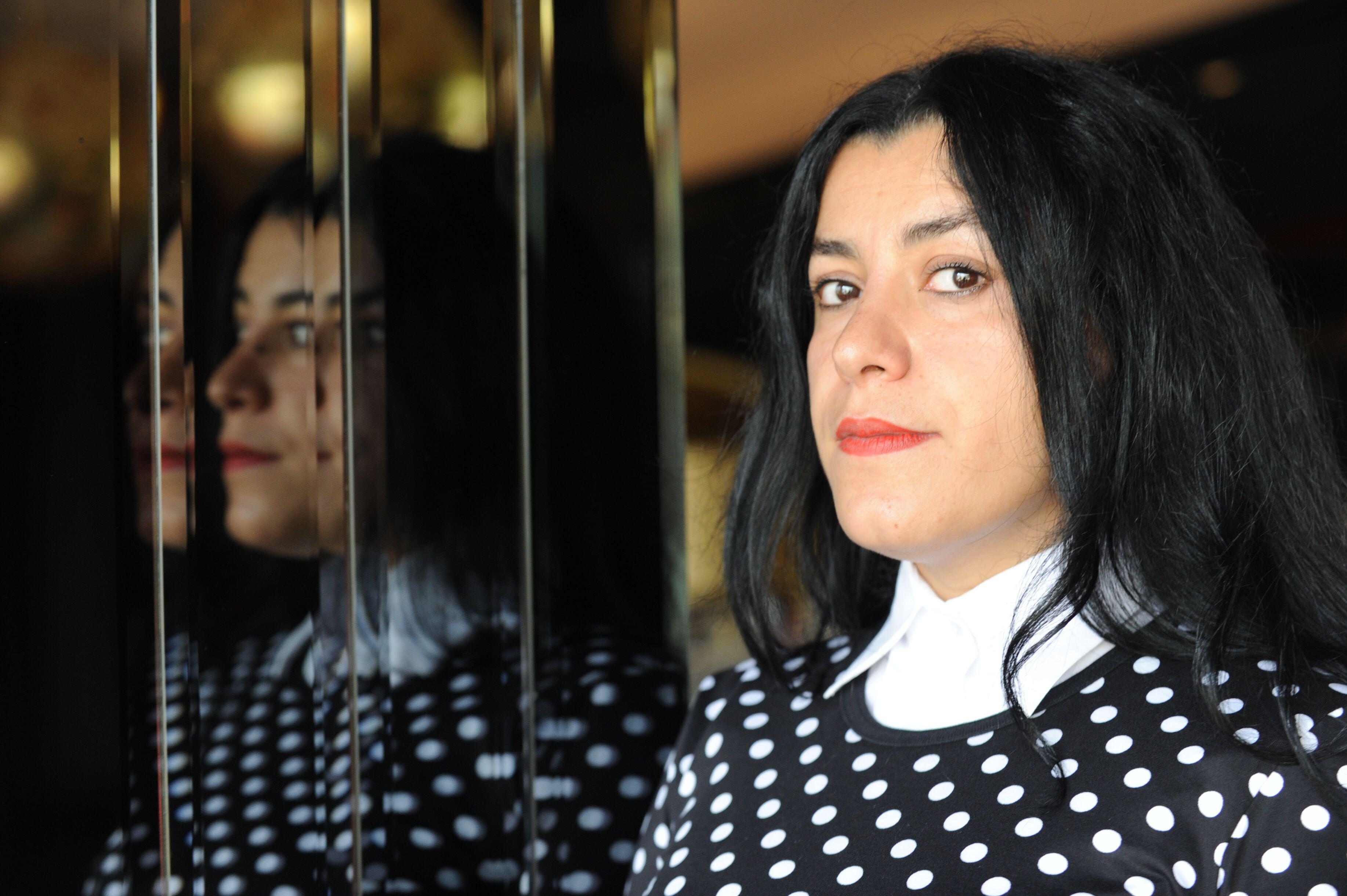 Photo of Marjane Satrapi dressed in a white spotted gown with a collared shirt, her reflection is on the left.