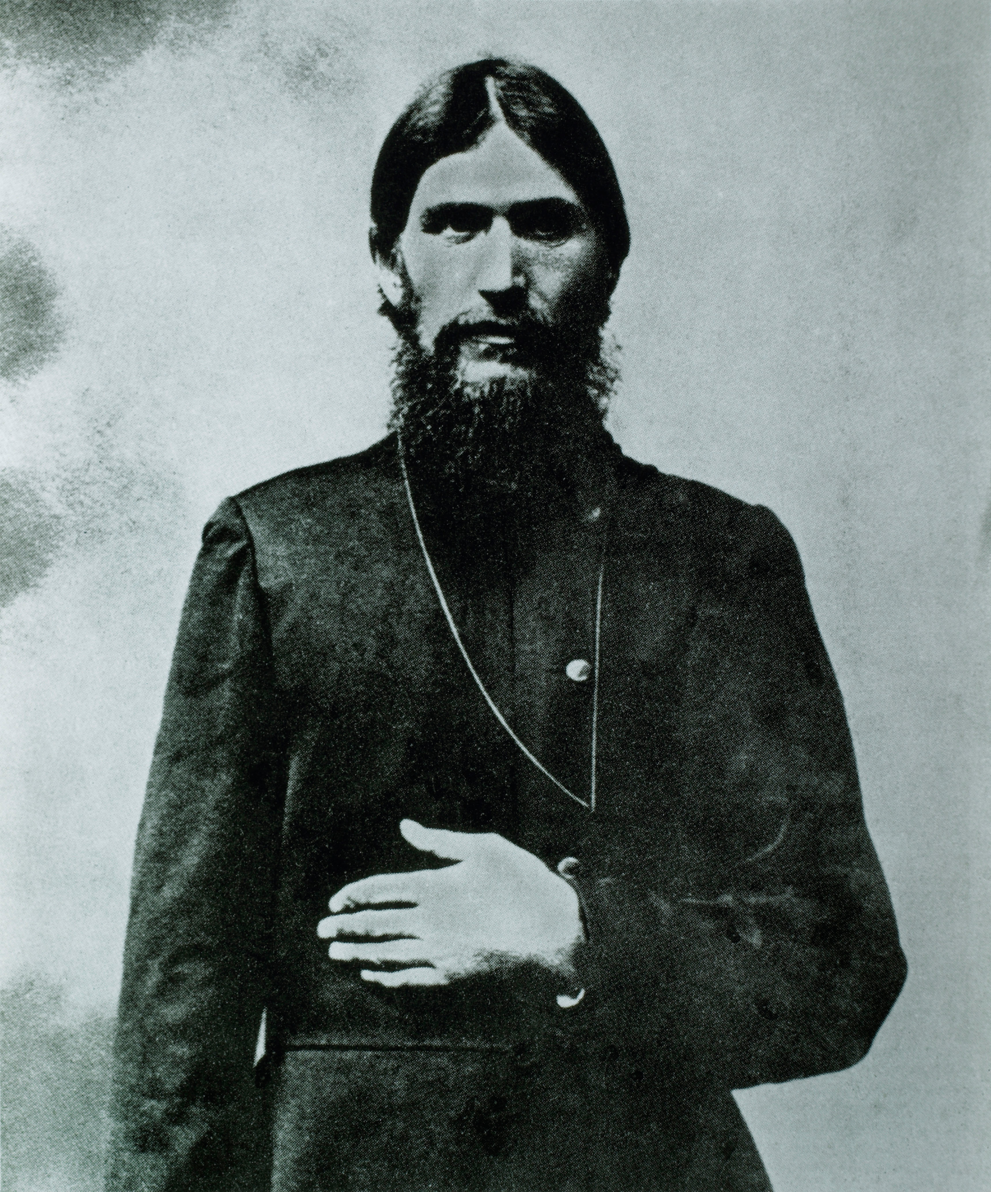 A photo of the real Rasputin, dressed in black, his left hand is resting on his stomach