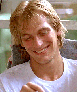 A young Julian Sands smiles  while sitting in a white t-shirt