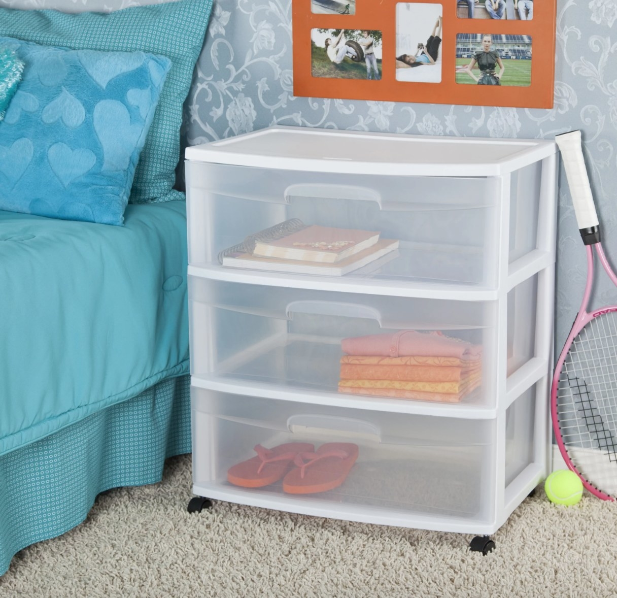 a clear plastic rolling cart with three drawers filled with folded shirts, a pair of flip flops, and books in a bedroom