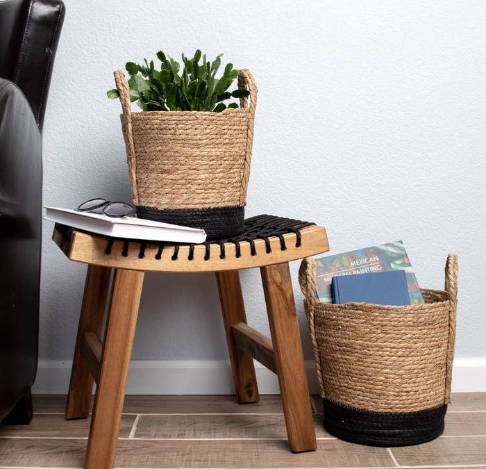 a set of two different-sized rattan baskets with books and plants in them, one basket is sitting on a wooden stool