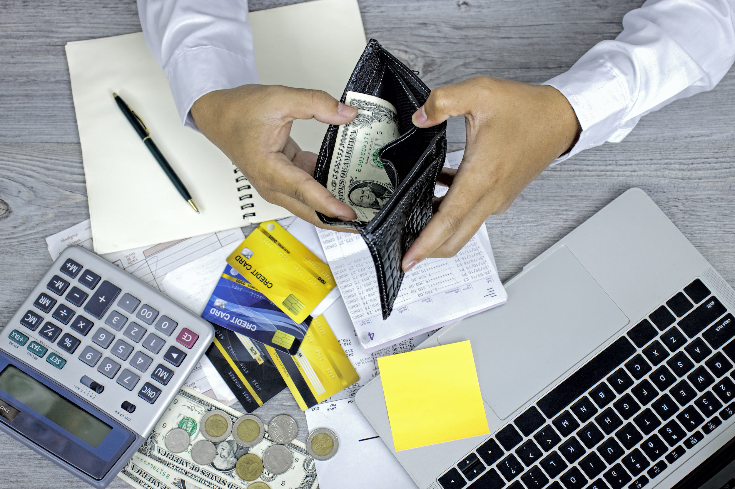 A person holding an open billfold above a desktop with a laptop, calculator, credit cards, bank statement, and cash
