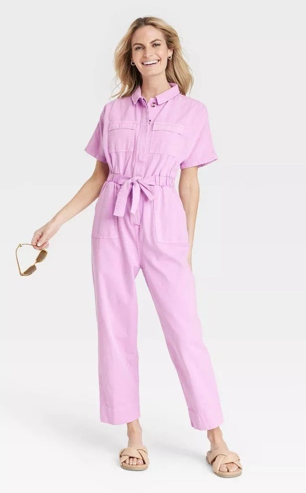 A smiling model wearing the boilersuit in pink while holding a pair of sunglasses