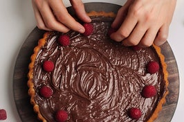 a pair of hands arranging raspberries on top of a chocolate tart
