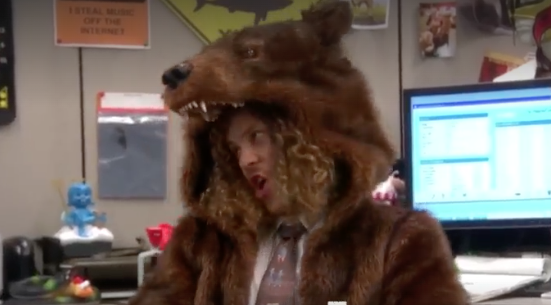 Woman in office wears bear costume with open jaw headpiece; background monitors, figurines, and posters visible