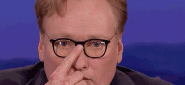 Conan O&#x27;Brien is touching his nose, looking pensive, with a blurred blue backdrop