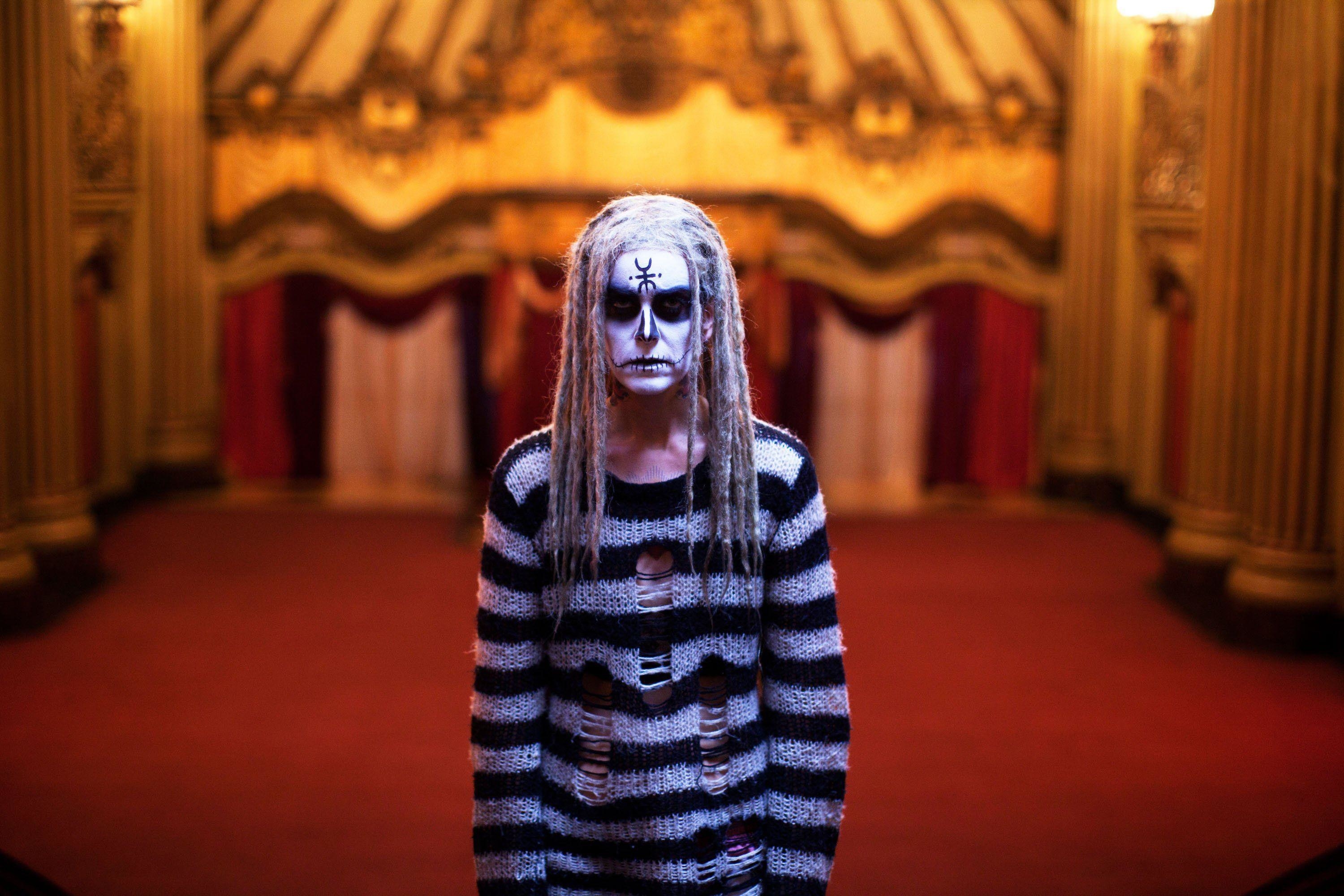 A woman in creepy make-up and a long worn-out sweater stands in an ornate ballroom