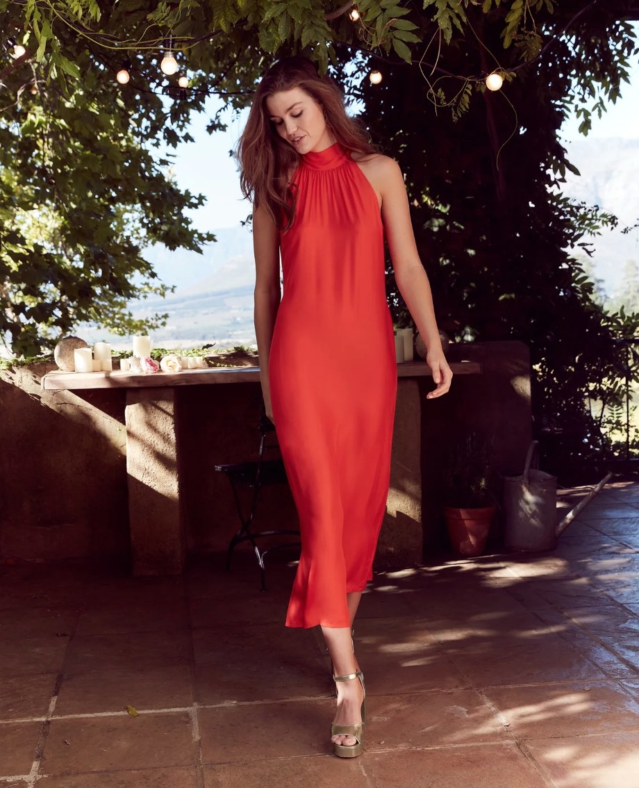 a model on a countryside wearing a red satin midi dress with a halter neck and gold platform sandals