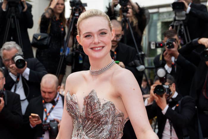 A closeup of Elle smiling as she poses in her sequined strapless outfit at a red carpet event with paparazzi behind her