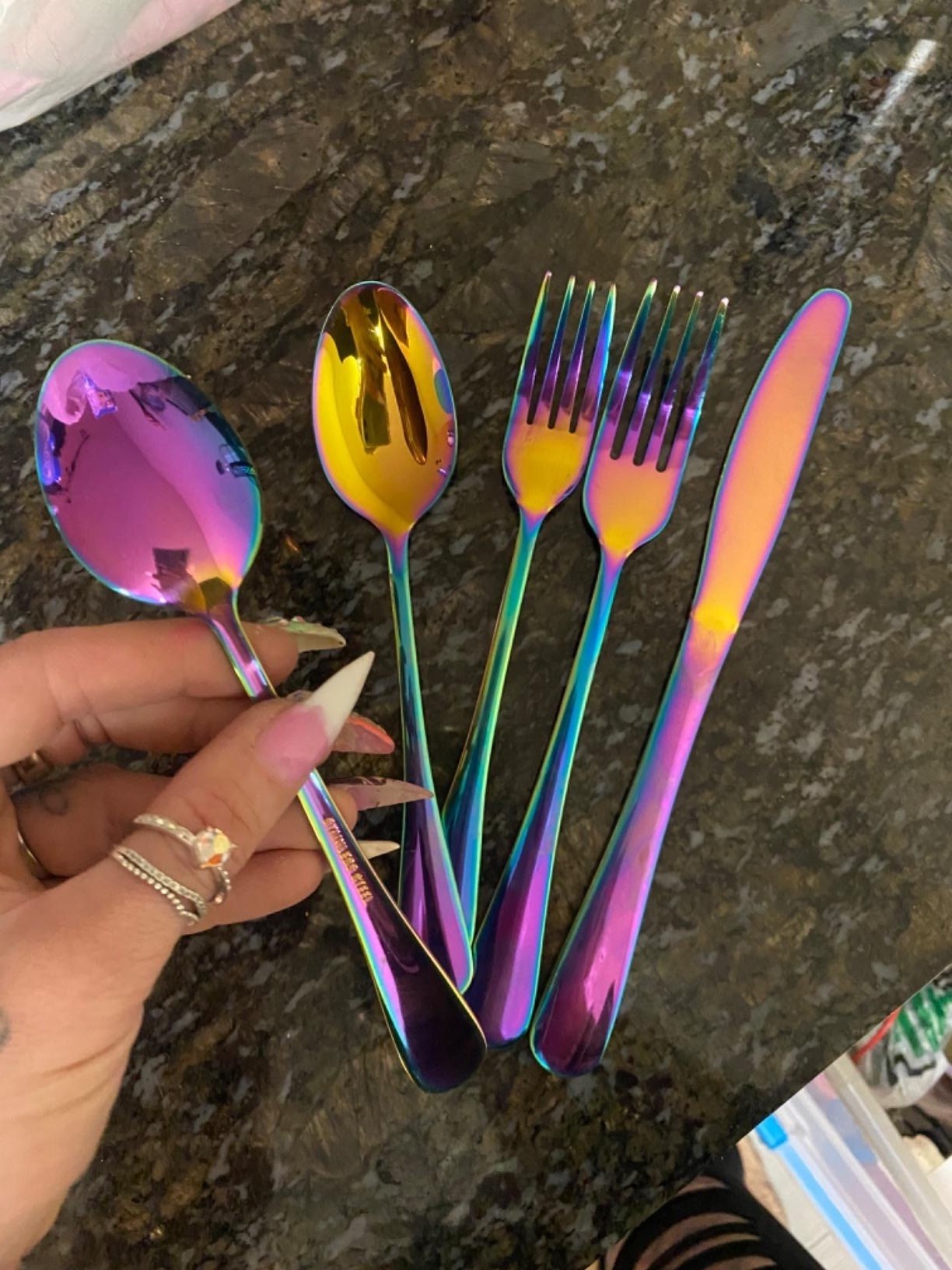 a reviewer holding up a rainbow spoon while four other rainbow utensils lay on a countertop