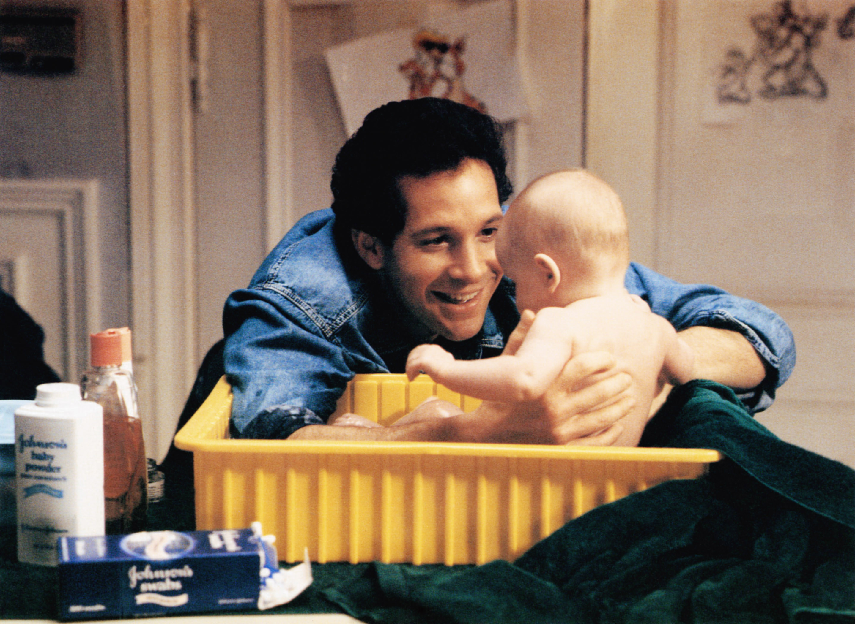 Steve Guttenberg holding a baby, played by twins Lisa and Michelle Blair, in a yellow bucket in the movie Three Men and a Baby