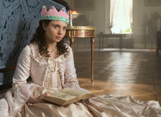 Young Violet sits on the ground wearing a paper crown and reading