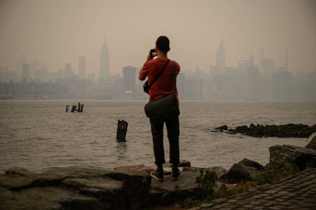 A man stands on rocks alongside the East River and takes photos of the Manhattan skyline, shrouded in smoke