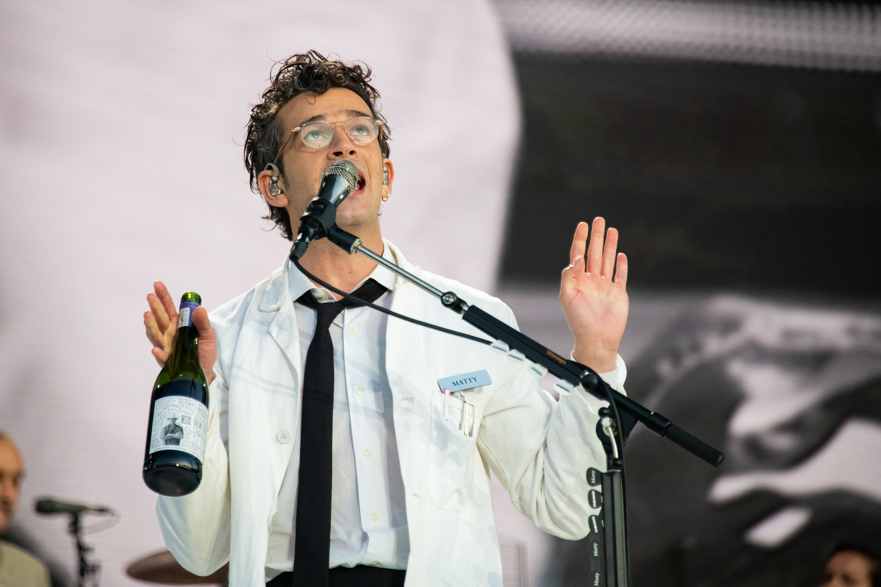 Matt holding a bottle of wine during a performance in which his wearing what looks like a lab coat and a name tag