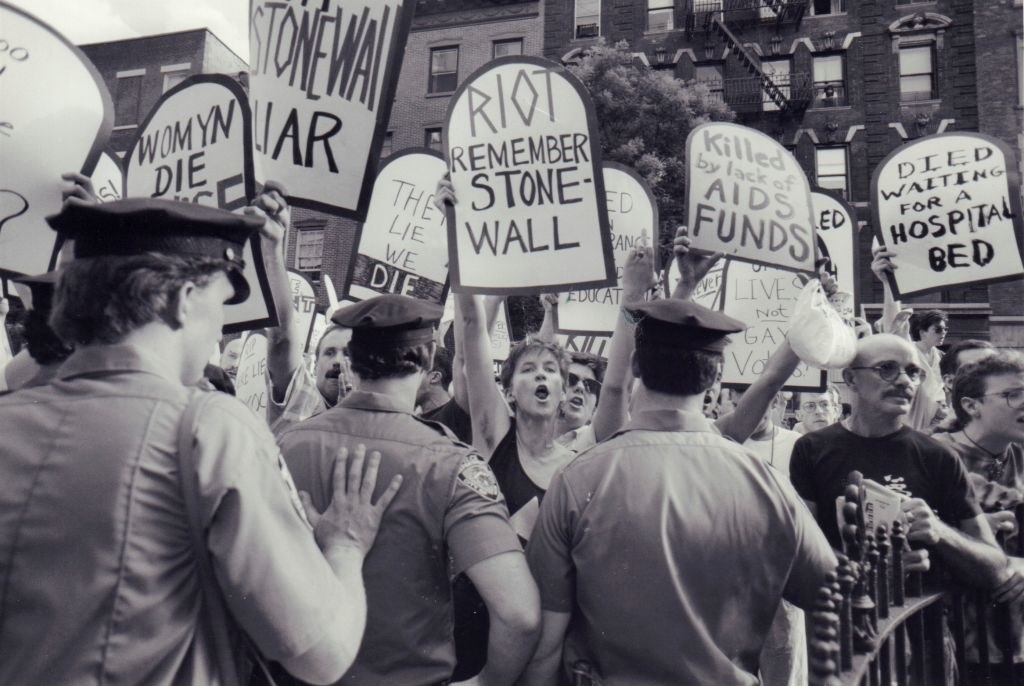 A black-and-white photo of a protesters with signs. Some signs say &quot;RIOT REMEMBER STONEWALL&quot; and &quot;Killed by lack of AIDS Funds&quot;