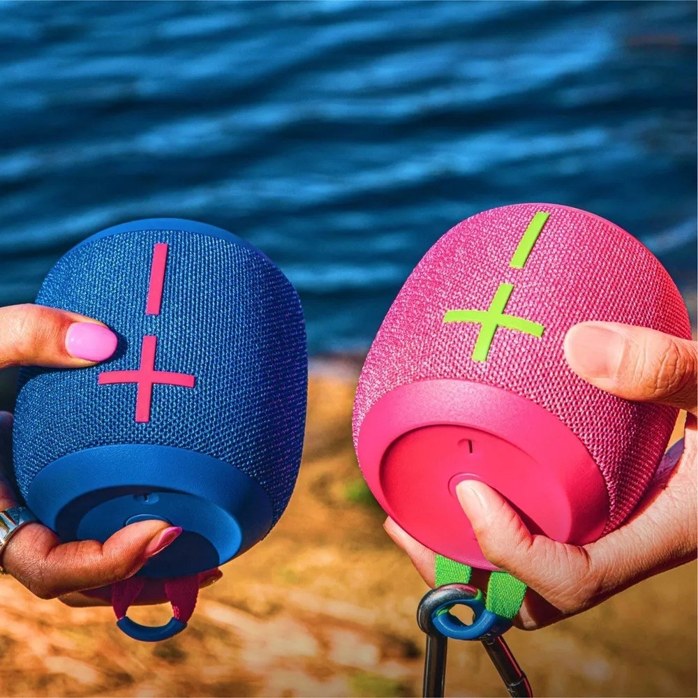 Two hands holding portable wireless speakers near a body of water