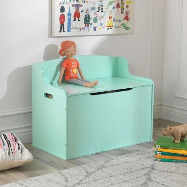 a mint-colored toy box with attached seat holding a doll