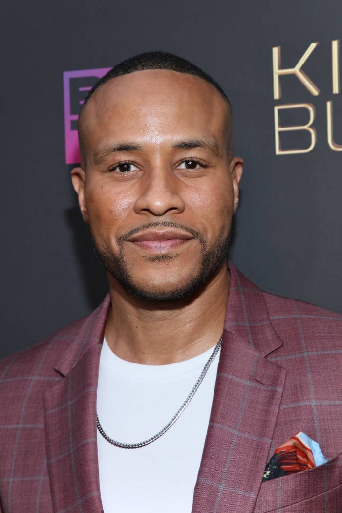 A close-up of DeVon Franklin at a red carpet event; he is wearing a plaid jacket with a pocket square, a simple T-shirt, and a thin chain