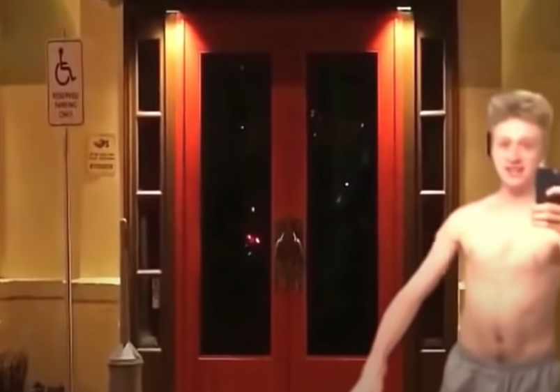 A shirtless young man taking a selfie in front of entrance doors