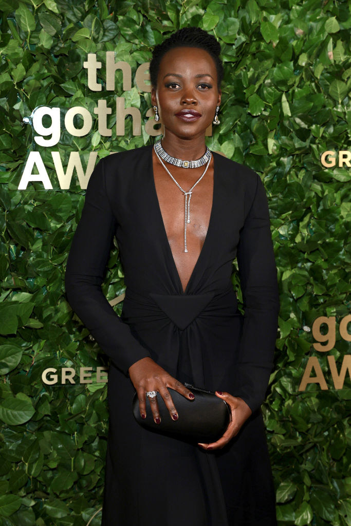 Lupita in a long-sleeved, deep-V outfit and standing in front of a leafy background