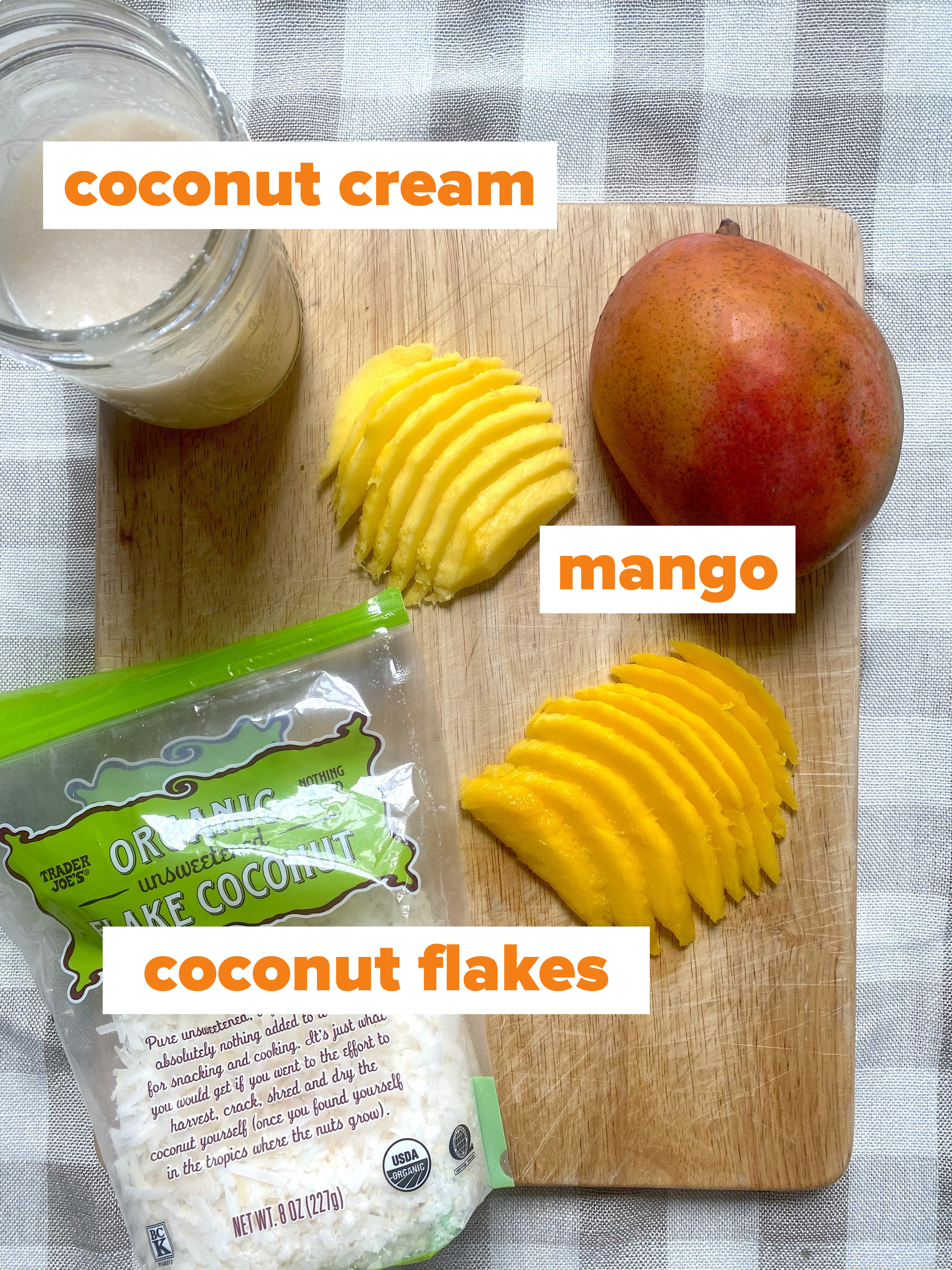 A cutting board with a jar of coconut cream, sliced mango, and a bag of coconut flakes