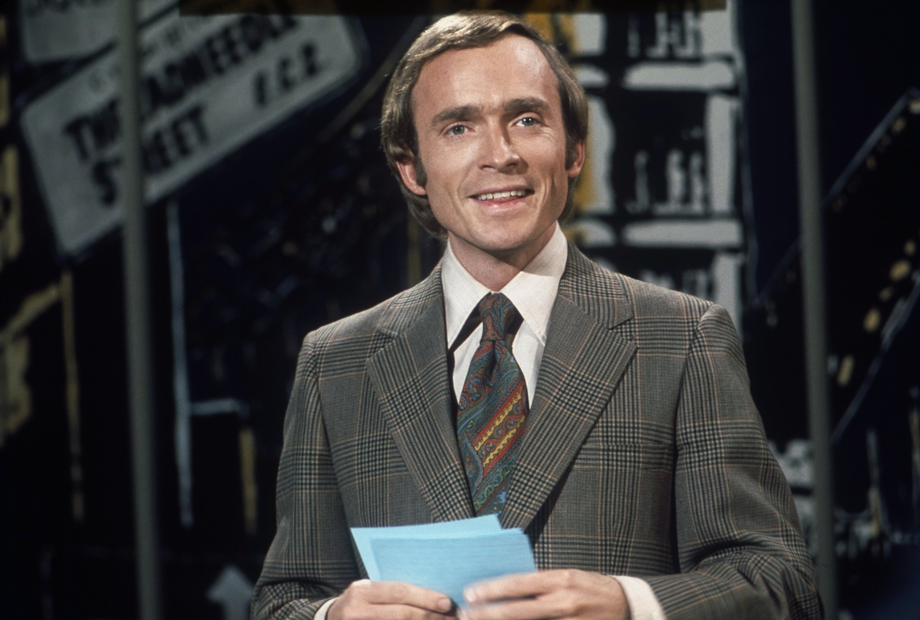 Cavett holding sheets of paper and wearing a suit and tie