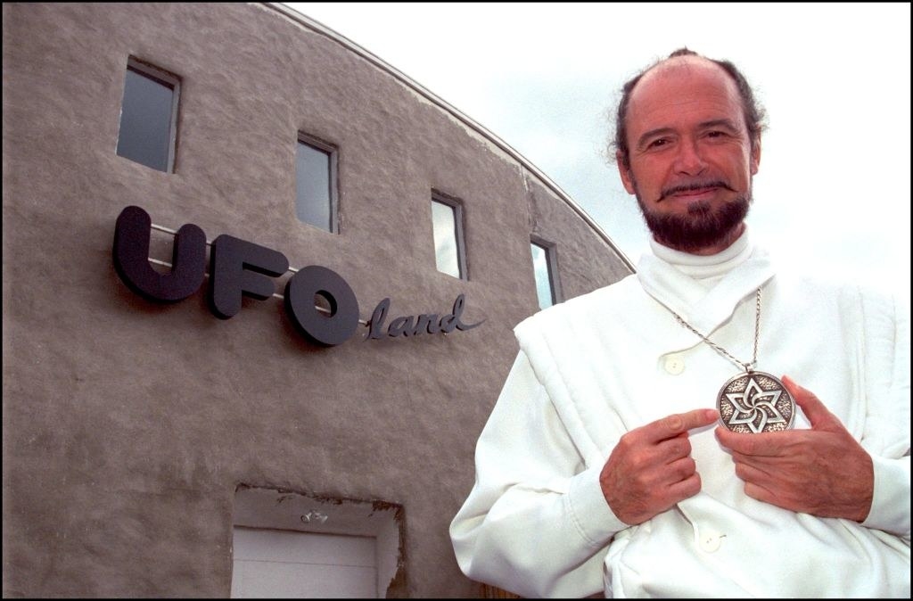 a man wearing a pointing to his pendant necklace while standing outside of a building called ufo land