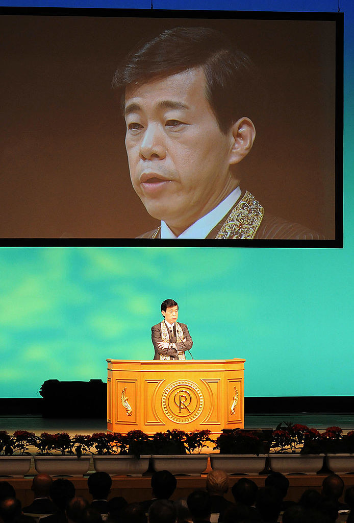 man speaking at a podium with a screen above him projecting his face