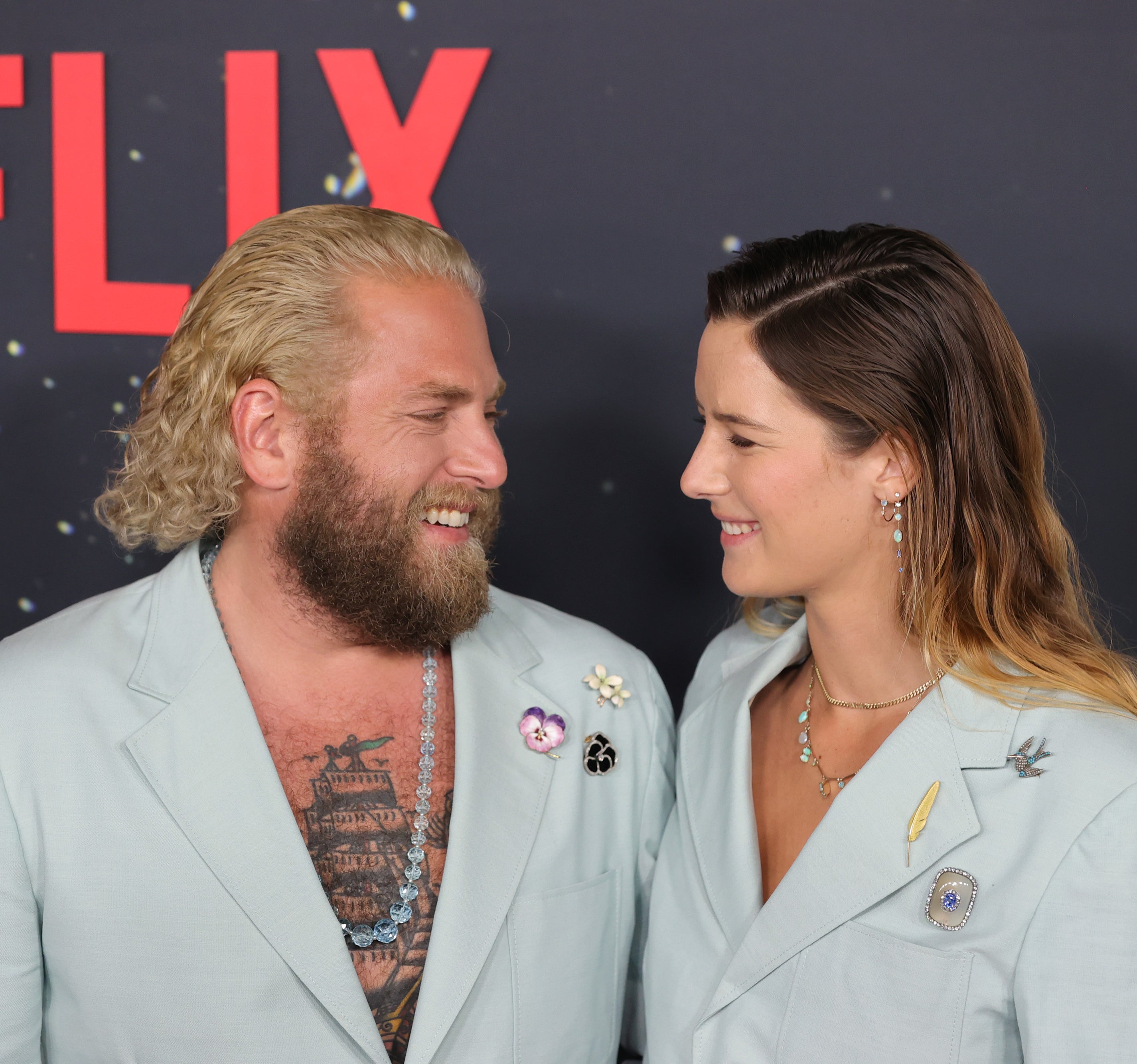 Close-up of Jonah and Sarah at a press event wearing matching suits and smiling at each other