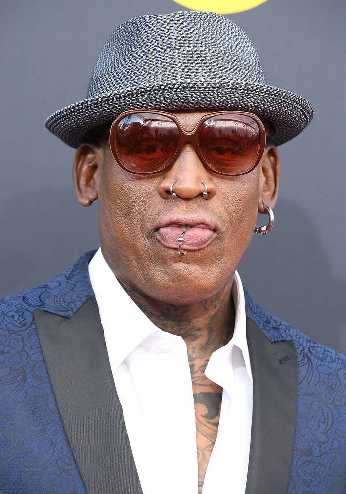 A close-up of Dennis Rodman in an open suit, sunglasses, and a trilby hat
