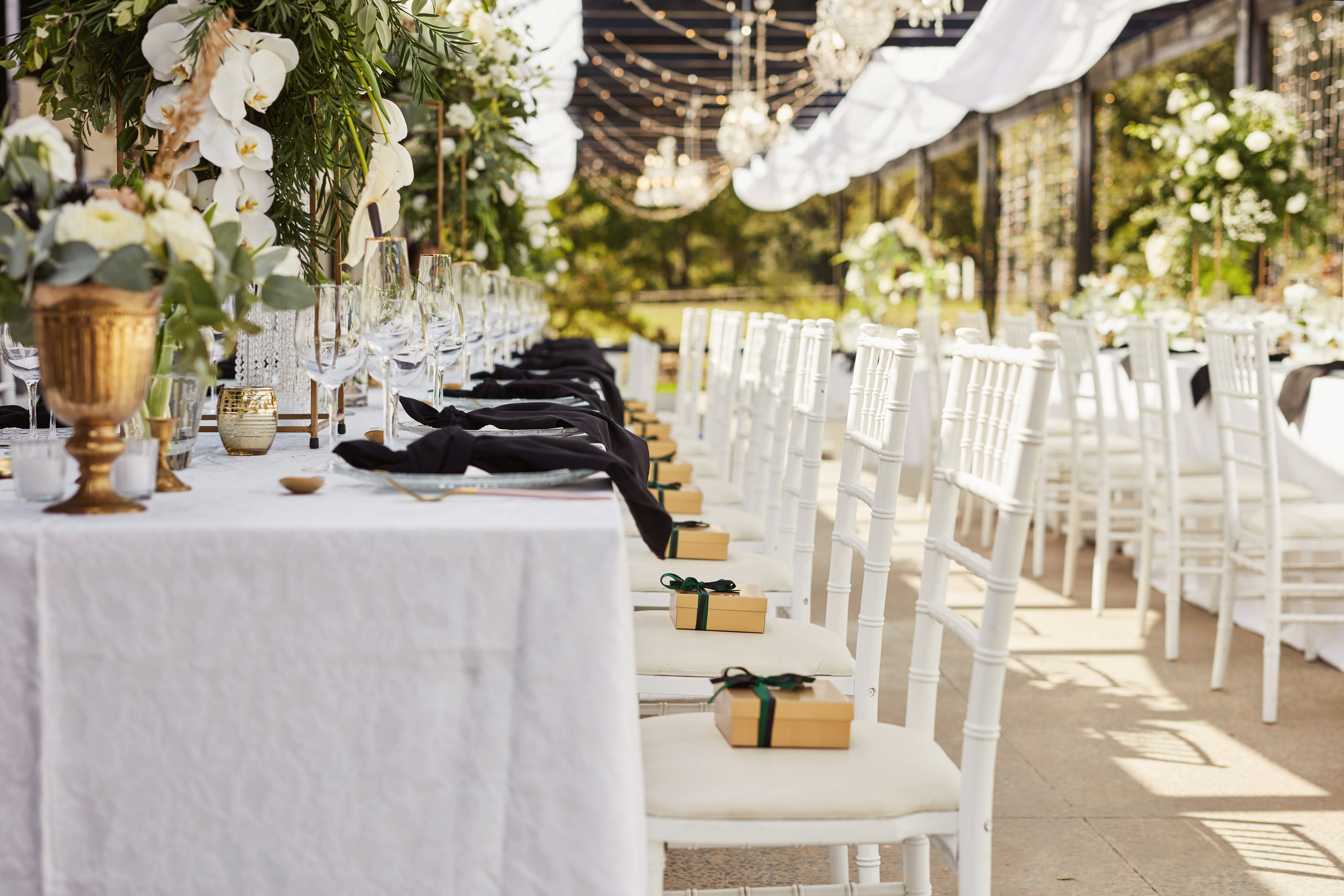 A wedding reception with lush green centerpieces, gold accents, white chairs, and small boxes with black ribbon as guest favors