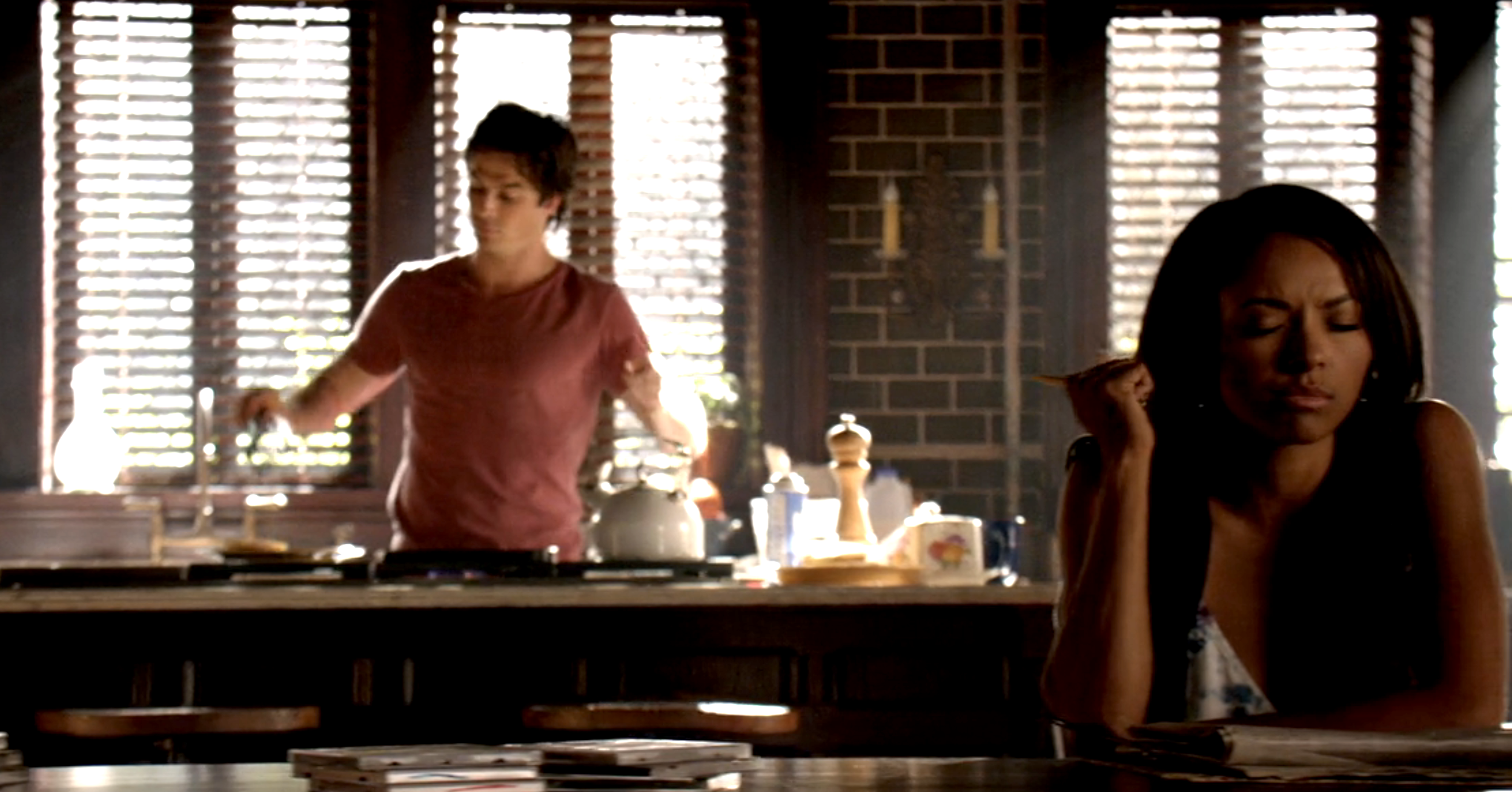 Damon making pancakes while Bonnie sits at the table thinking