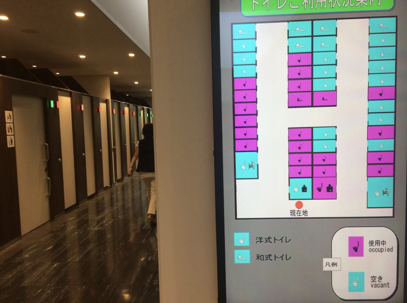 In front of a line of toilets is an electronic screen that shows the row of toilets and has icons indicating which are currently occupied