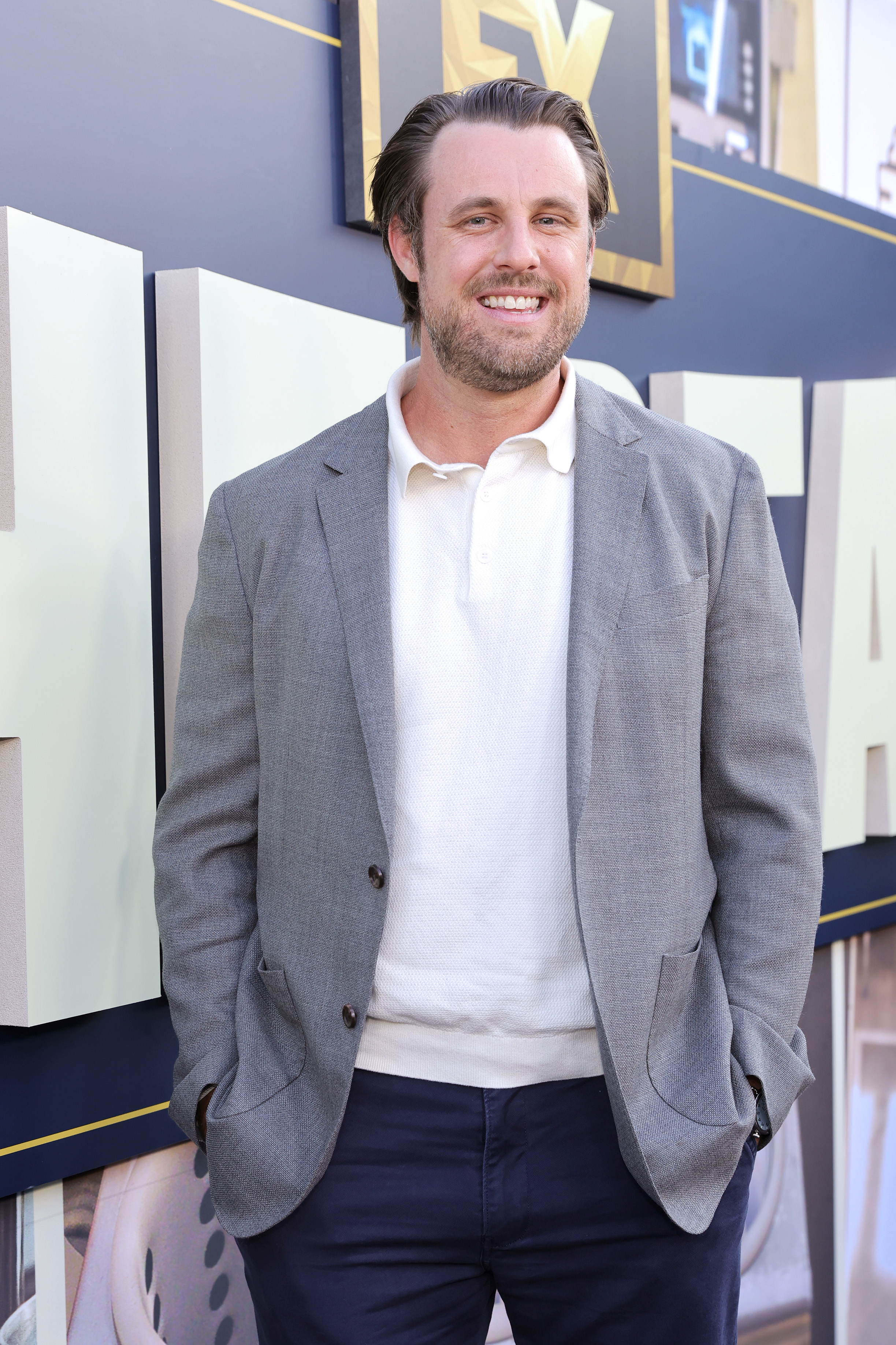 Chris Witaske on the red carpet wearing a blazer, polo short, and slacks. He smiles at the camera while he stands with his hands in his pockets