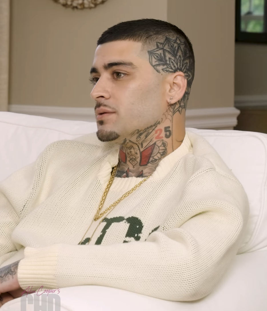 A closeup of Zayn sitting on a couch during his interview. Zayn is wearing a sweater and his hair is closely shaved on the sides showing off his head tattoo