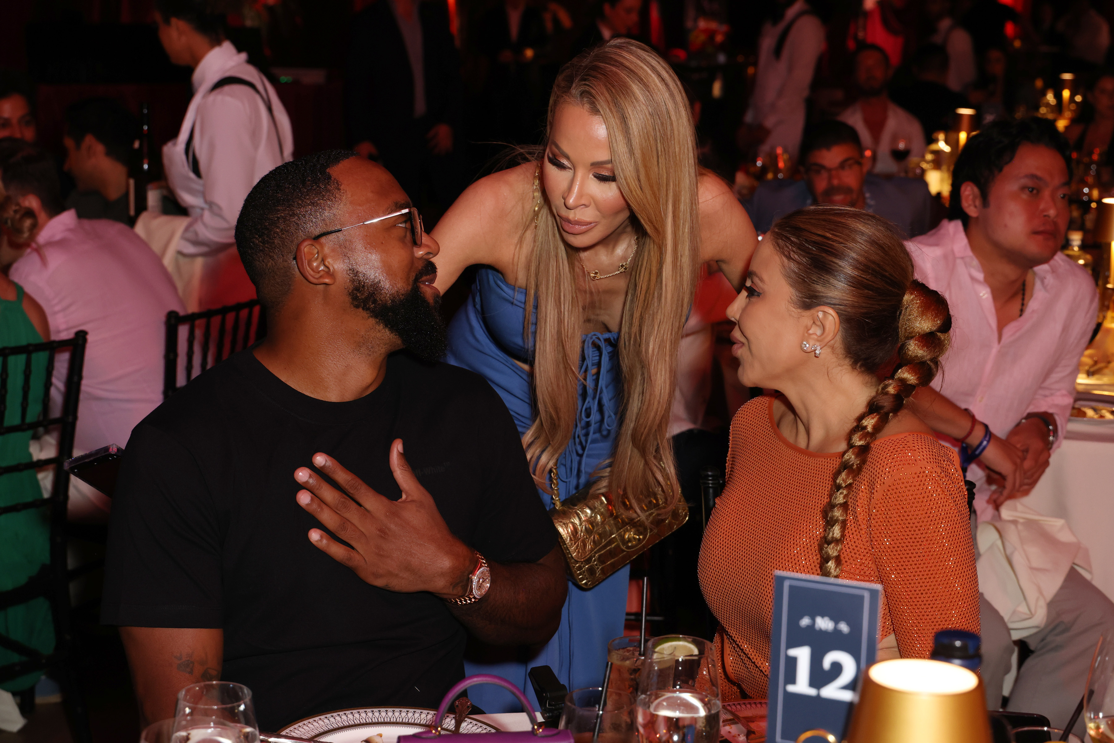 Lisa Hochstein leaning down to speak to Marcus and Larsa who are sitting at a round dinner during an event