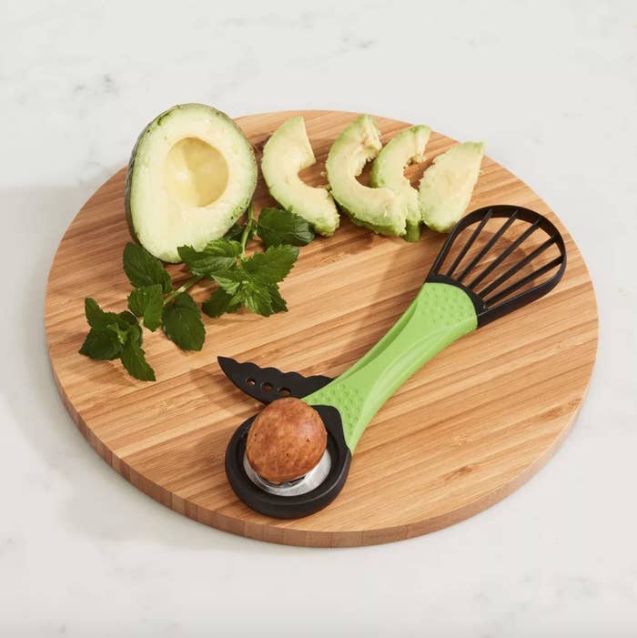 an avocado, the avocado cutting tool, and herbs on a round wooden cutting board