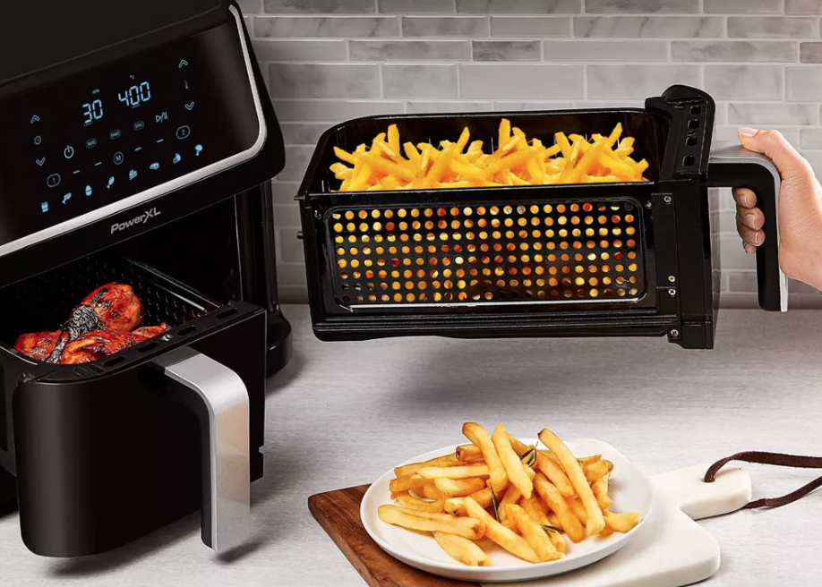 a person taking one side of the air fryer basket out that holds french fries, the other basket is open holding chicken. there&#x27;s a plate of french fries in the foreground