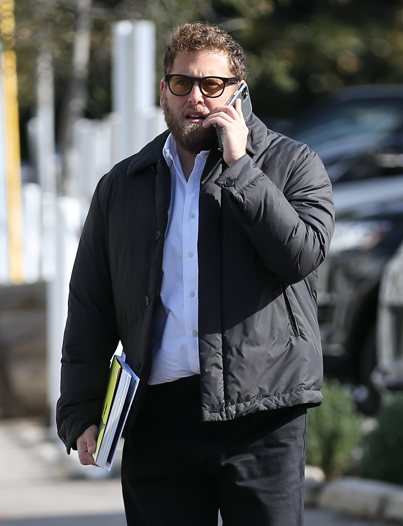 Jonah Hill walking outside while talking on the phone and holding a stack of papers