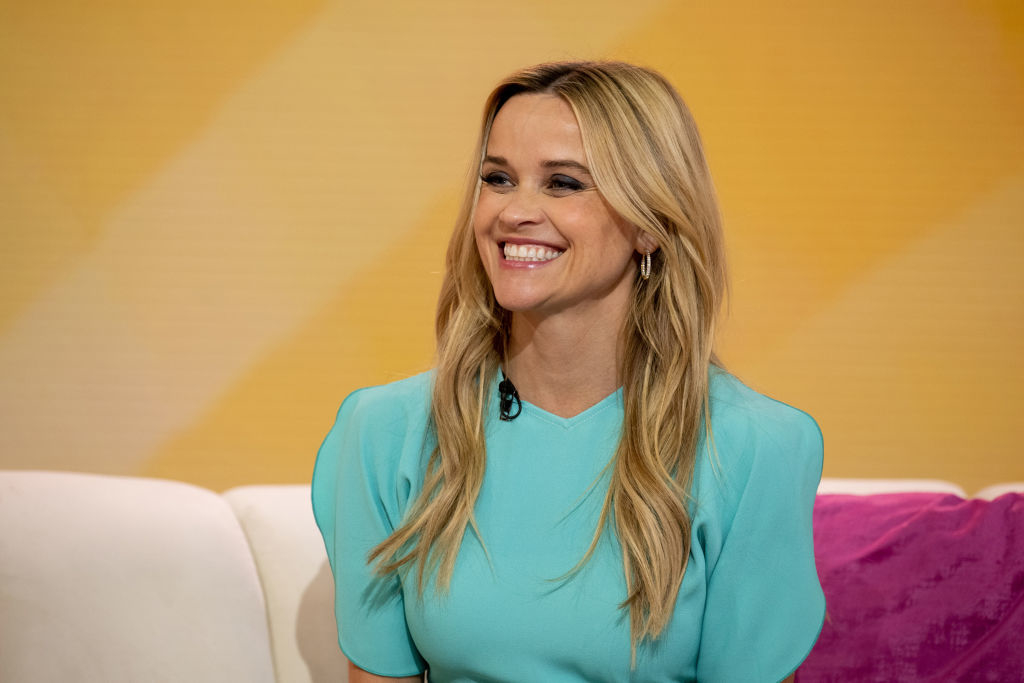 An older Reese smiles as she sits on a couch for an interview