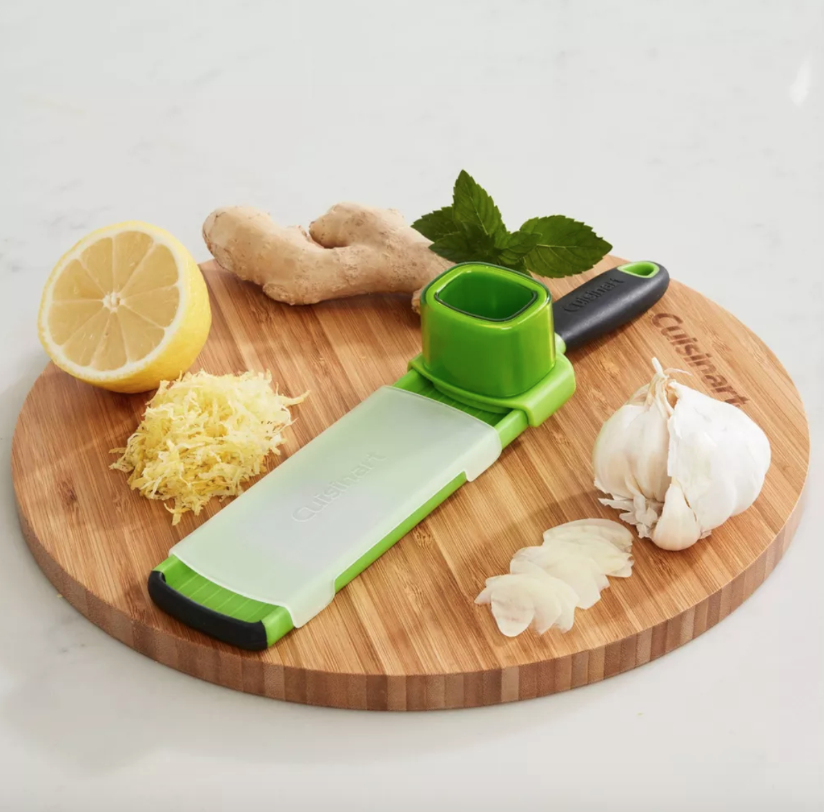 a garlic slicer tool, a lemon, lemon zest, garlic, ginger, and basil on a round wooden cutting board