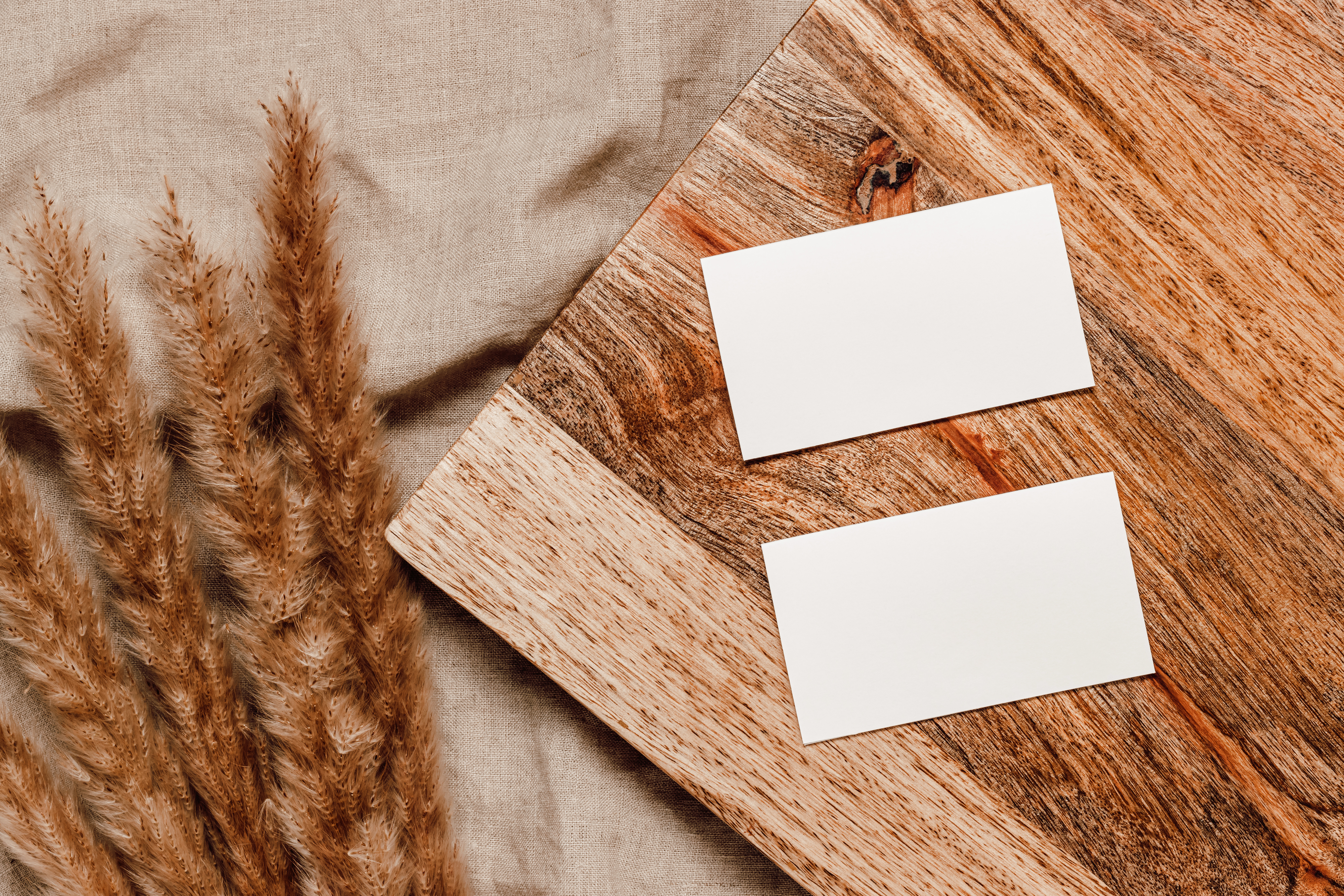 Two blank white cards, the size of credit cards, sit on a piece of dark wood. The wood is on top of a beige sheet with a bunch of dried wheat-like flowers next to it.