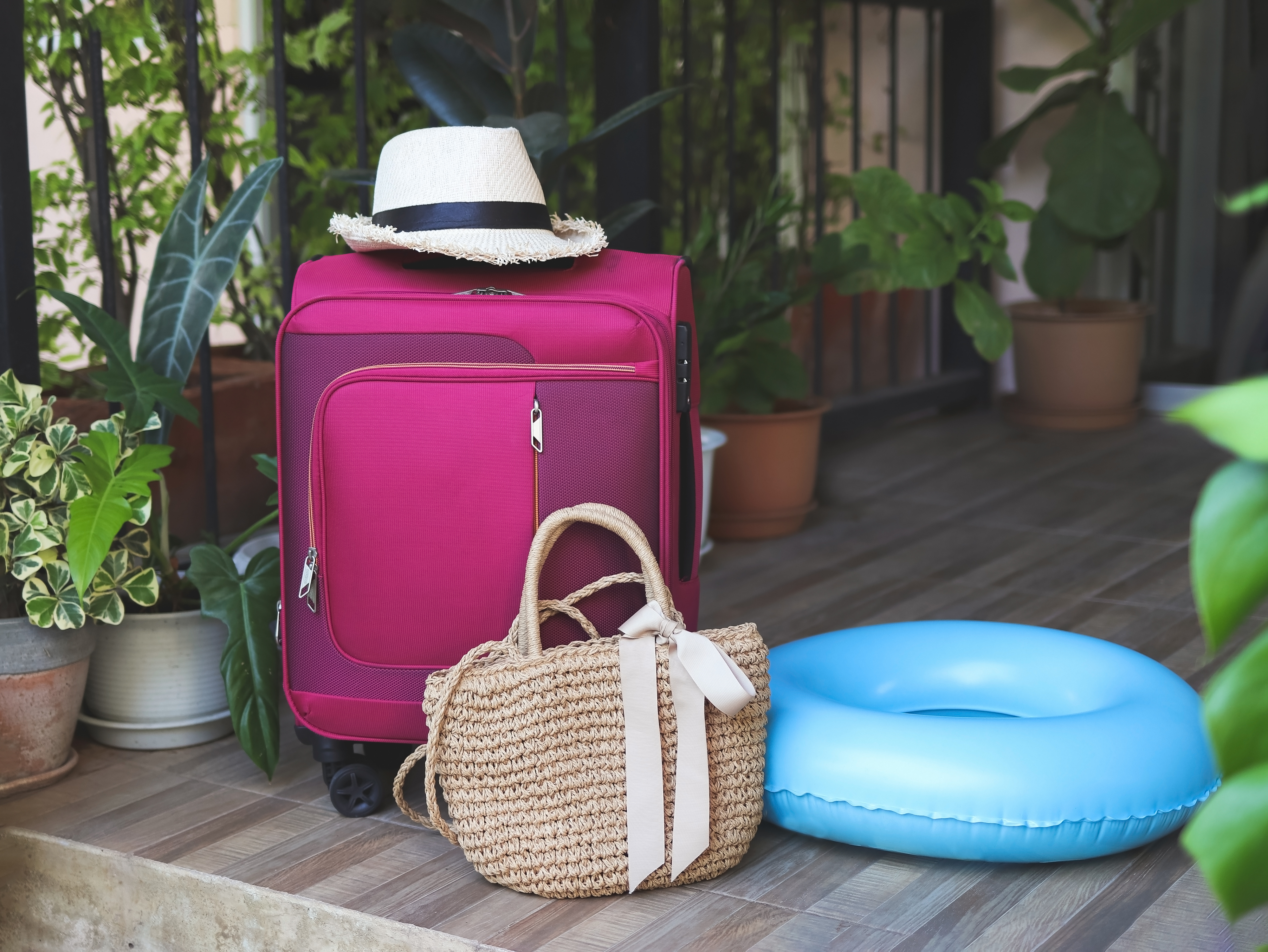 An image of a pink and plum colored suitcase, with a hessian bag and a blue rubber ring next to it. There is a straw hat on top of the suitcase.
