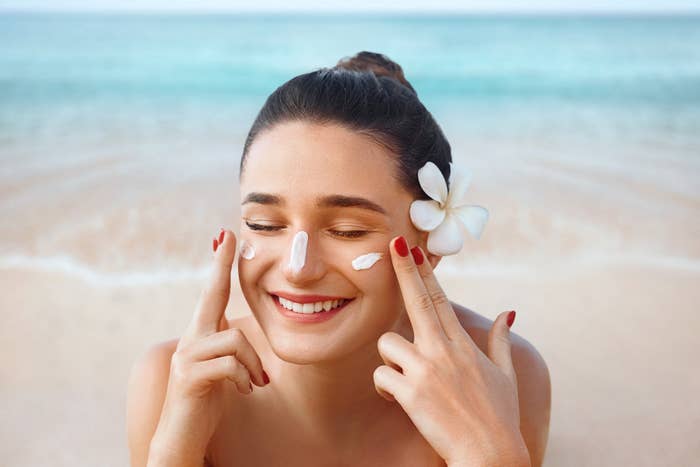 A woman applying sunscreen to her face. She has a frangipani flower on the left side of her head, and is smiling with her eyes shut.