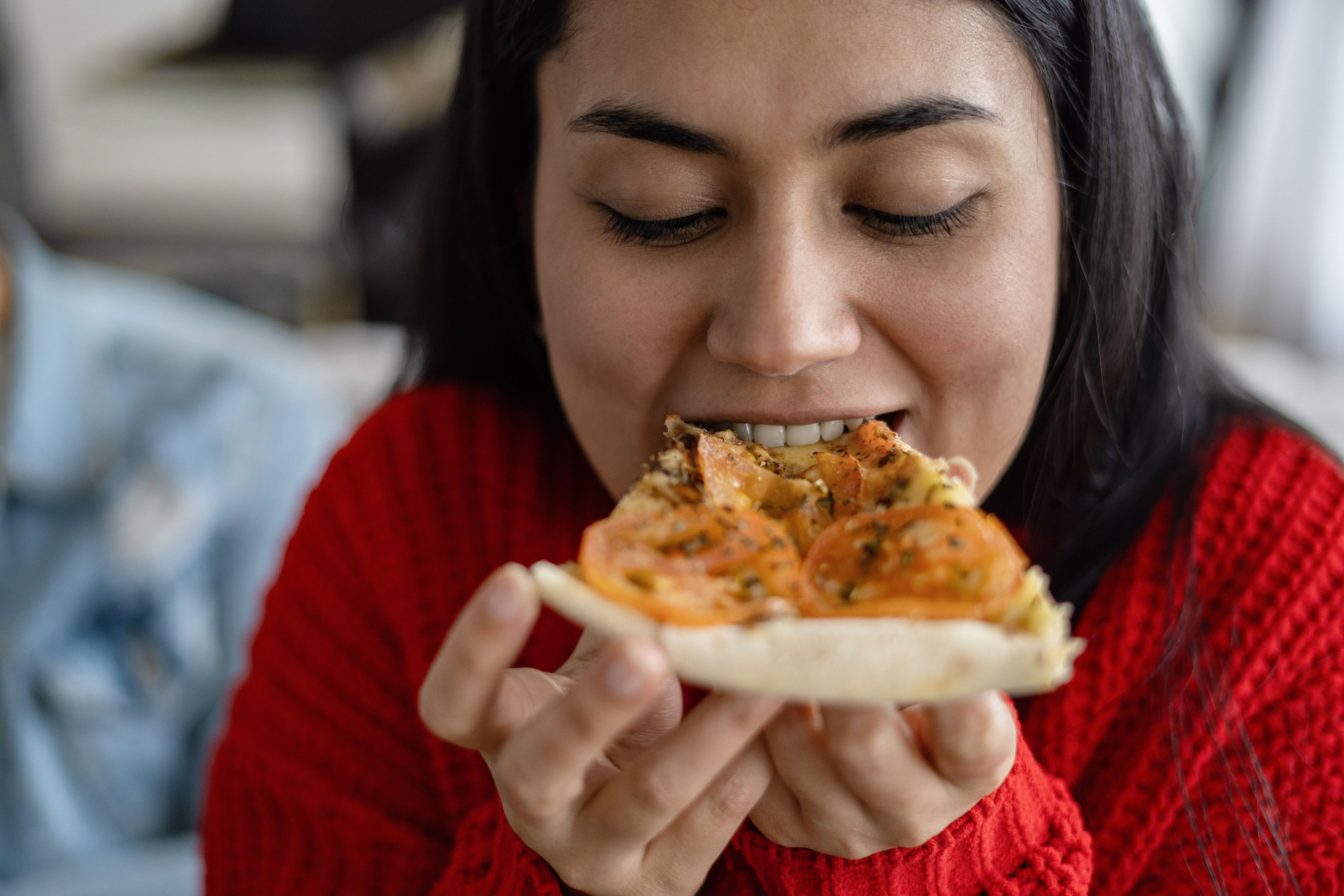 A woman biting a slice of pizza. She has dark hair and is wearing a red knitted sweater. She is looking down as she bites into it.