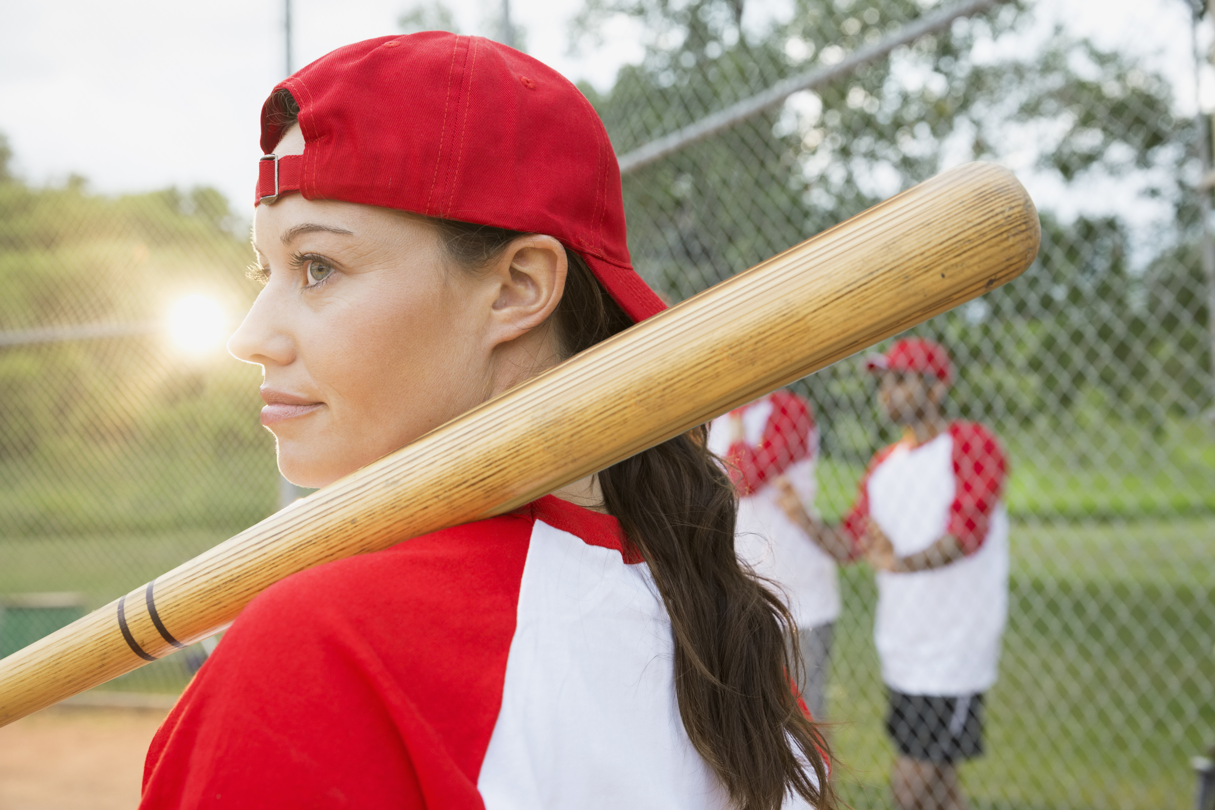An image of a woman playing baseball. She&#x27;s wearing a red cap backwards and has the bat resting on her shoulder.