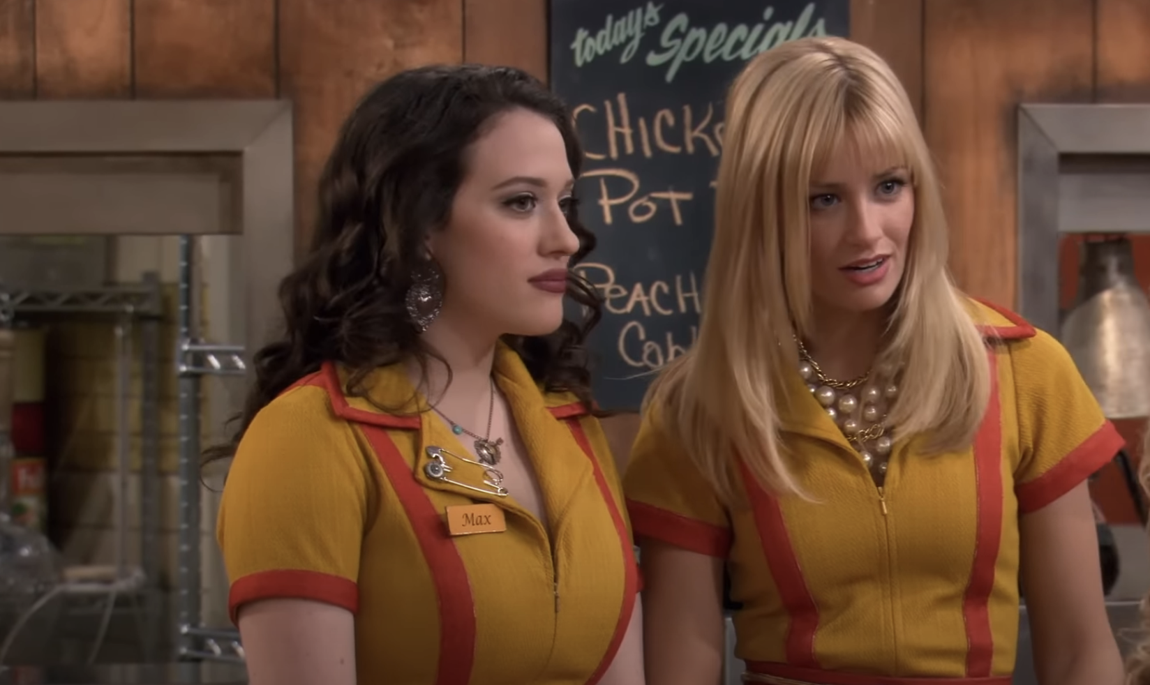 Two women in diner uniforms stand together. One has dark, wavy hair and the other has straight, blonde hair. A menu board is visible behind them